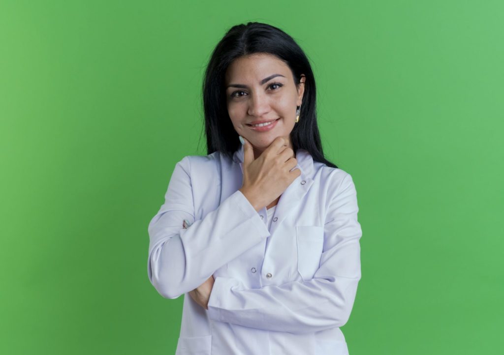 Smiling Young Female Doctor Wearing Medical Robe Looking At Camera Touching Chin Isolated On Green Background With Copy Space